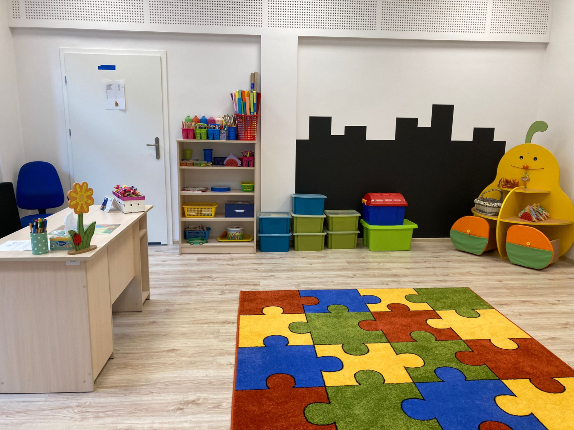 Sala gr. pierwszej. Po lewej stronie znajduje się biurko nauczycielki. Na podłodze leży kolorowy dywan. Na wprost znajduje się regał oraz pojemniki na zabawki. Po prawej stronie jest regał w kształcie gruszki.