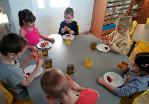 Dzieci siedzą przy stolikach i jedzą pizzę.