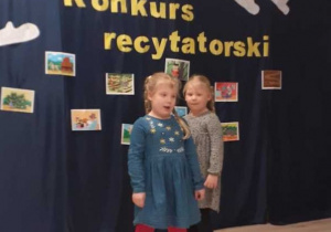 Dziewczynki recytują wiersz. Za nimi znajduje się dekoracja: napis Konkurs recytatorski oraz ilustracje do wierszy/