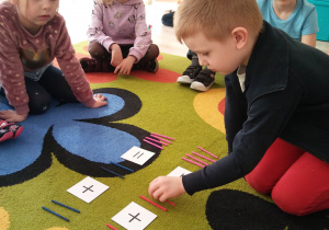 Dzieci siedzą w kole i uczestniczą w zabawie matematycznej z wykorzystaniem patyczków.