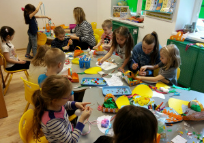 Dzieci siedzą przy stolikach wraz z dwoma mamami i wykonują wielkanocne prace plastyczne. Na stolikach leżą między innymi: kolorowe piórka, szablony kur, styropianowe jajka, cekiny, pompony, itd. Z tyłu siedzi, przy stoliku, siedzi pani nauczycielka, która pomaga dziewczynce odciąć kawałek wstążki.