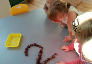 Dziewczynki i chłopiec siedzą przy stoliku. Układają z kasztanów kształt cyfry 2.