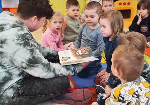 Dzieci siedzą w kole na dywanie. Wśród nich siedzi chłopak, który trzyma książkę i czyta na głos dzieciom bajki.