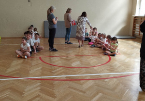 Dzieci siedzą w dwóch rzędach. Pani dyrektor szkoły rozdaje dzieciom słodkie upominki.