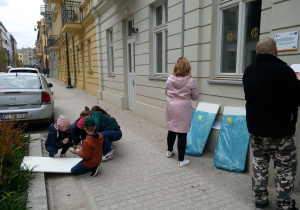 Ulica Włókiennicza. Uczestnicy malują obraz na płótnie położonym na chodniku. Obok stoi pani nauczycielka koordynująca grę.