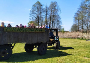 Dzieci, wraz z nauczycielkami, jadą drewnianą przyczepą ciągniętą przez traktor.