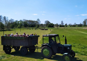 Dzieci, wraz z nauczycielkami, jadą drewnianą przyczepą ciągniętą przez traktor.