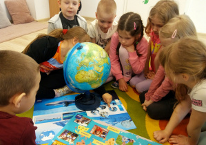 Dzieci siedzą w kole na dywanie. Przed nimi stoi globus, a na podłodze leżą plansze edukacyjne przedstawiające kontynenty i zwierzęta egzotyczne.