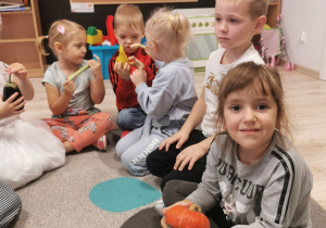 Na zdjęciu znajdują się dzieci siedzące na dywanie. Dziewczynka na pierwszym planie trzyma w rękach dynię. Dzieci na drugim planie oglądają pozostałe warzywa.