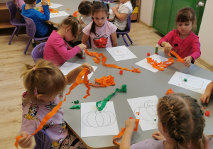 Na zdjęciu na pierwszym i drugim planie widoczne są stoliki w sali przedszkolnej, przy których siedzą dzieci. Dzieci wykonują pracę plastyczną przy użyciu pasków z bibuły. Na stolikach leżą kartki.