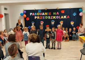 Na zdjęciu widoczna jest sala gimnastyczna. Na pierwszym planie widnieją osoby dorosłe siedzące tyłem. Na drugim planie znajdują się dzieci ustawione w rzędzie, twarzami zwrócone przed siebie. Na trzecim planie widoczne są dzieci siedzące na ławce. Na czwartym planie widnieje dekoracja przedszkolna.