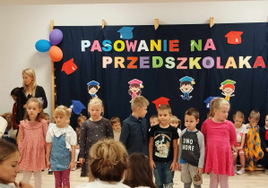 Na zdjęciu widoczna jest sala gimnastyczna. Na pierwszym planie znajdują się dzieci ustawione w rzędzie, twarzami zwrócone przed siebie. Na drugim planie widoczne są dzieci siedzące na ławce. Na trzecim planie widnieje dekoracja przedszkolna.