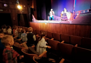 Na pierwszym planie dzieci tańczą na widowni w teatrze. W tle znajduje się scena z dekoracją statku.