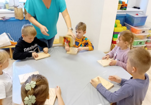 Na zdjęciu widoczne są dzieci siedzące przy stole. Dzieci wykonują pracę plastyczną przy pomocy sznurka. Na drugim planie widoczna jest nauczycielka prowadząca warsztaty plastyczne.