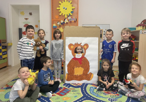 Na zdjęciu widoczna jest sala przedszkolna. W sali znajdują się dzieci. Niektóre dzieci siedzą na dywanie, inne, tuż za nimi stoją. blisko siebie. Twarze dzieci skierowane są w stronę aparatu. Dwoje dzieci stojących w środku trzyma podobiznę misia, tzw. fotobudkę. Na drugim planie znajduje się kaloryfer i drzwi z dekoracją przedszkolną.