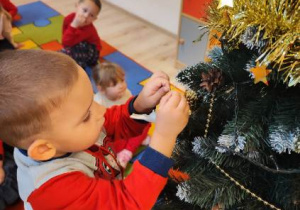 Zdjęcie wykonane zostało w sali przedszkolnej. Na pierwszym planie po lewej stronie widoczny jest chłopiec stojący bokiem do obiektywu, po jego prawej stronie widoczna jest choinka. Dziecko ubiera choinkę.