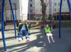 Dwie dziewczynki w kamizelkach odblaskowych bujają się na huśtawkach.