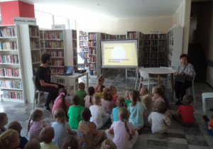 Dzieci znajdują się w bibliotece i oglądają prezentację wyświetlaną na projektorze. W tle widać regały z książkami.