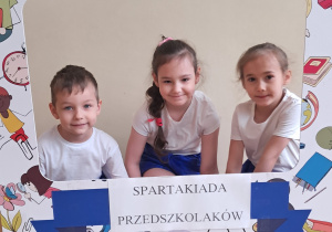 Chłopiec i dwie dziewczynki pozują do zdjęć z szablonem, na którym widnieje napis Spartakiada Przedszkolaków.