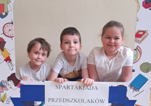 Dziewczynka, chłopiec i dziewczynka pozują do zdjęć z szablonem, na którym widnieje napis Spartakiada Przedszkolaków.