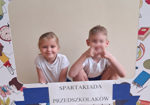 Dziewczynka i chłopiec pozują do zdjęć z szablonem, na którym widnieje napis Spartakiada Przedszkolaków.
