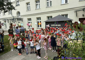 Na zdjęciu widać dzieci z całego przedszkola, które wspólnie śpiewają piosenkę i trzymają w ręku czerwone serduszka.