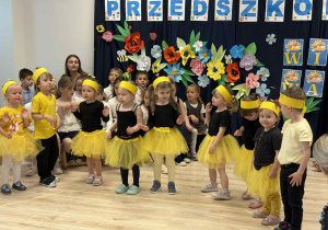 Na zdjęcu widać dzieci z najmłodszej grupy, które przebrane są w stroje pszczółek - żółte spódniczki i czarne bluzki. Dzieci śpiewają i naśladują pszczoły. Na zdjęciu widać także Panie Nauczycielki z naszego przedszkola.