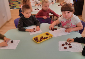 Dziewczynka i chłopcy siedzą przy stoliku, na którym leżą karty pracy dotyczące drzew. Na kartach pracy oraz na środku stolika leżą kasztany.