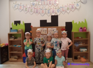 Ósemka dzieci pozuje do zdjęcia w koronach, stojąc pod napisem Dzień Przedszkolaka.