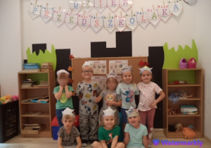 Ósemka dzieci pozuje do zdjęcia w koronach, stojąc pod napisem Dzień Przedszkolaka.