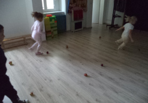 Sala gimnastyczna. Dwie dziewczynki i chłopiec biegną slalomem wokół jabłek.