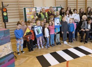 Grupa dzieci przedszkolnych, szkolnych oraz seniorów pozuje do zdjęcia na tle rekwizytów związanych z bezpieczeństwem w ruchu drogowym.