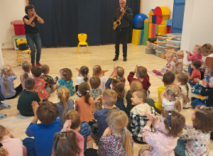 Dzieci siedzą na podłodze i klaszczą, podczas gdy muzyk gra na skasofonie.