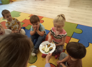 Dzieci siedzą na dywanie. Dziewczynka bierze z talerza owoc.