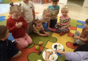 Dzieci siedzą na dywanie i obserwują jak nauczcyielka obiera owoce.