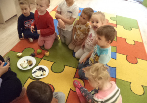 Dzieci siedzą na dywanie. Przed nimi, na talerzu, leżą kawałki owoców.