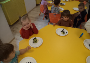Dzieci siedzą przy stołach. Przed nimi, na talerzach, leżą zrobione z owoców jeże.