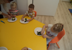 Dzieci siedzą przy stole i tworzą jeże z owoców. Pomaga im nauczyciel.