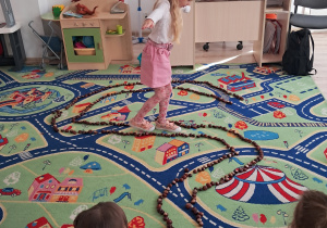 Dziewczynka idzie wąską ścieżką ułożoną z kasztanów. Dla utrzymania równowagi ma rozłożone na boki ręce.