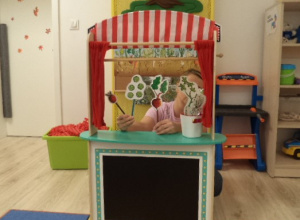 Na zdjęciu widać teatrzyk kukiełowy. Zza czerwonej kotary nauczycielka prezentuje dzieciom warzywne postacie.