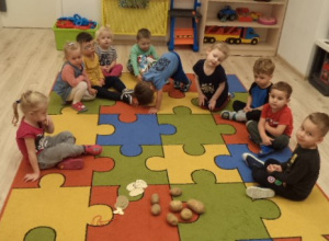 Grupa dzieci siedzi na dywanie. Przed nimi leżą ziemniaki i medale w kształcie ziemniaków.