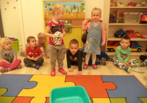 Grupa dzieci siedzi na dywanie. Dwie dziewczynki rzucają ziemniakiem do miski.