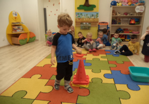 Grupa dzieci siedzi na dywanie. Chłopiec niesie na obręczy ziemniaka.