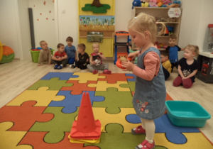 Grupa dzieci siedzi na dywanie. Dziewczynka niesie na obręczy ziemniaka.