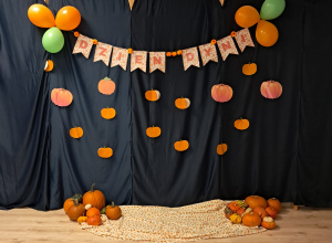 Dekoracja na dzień dyni. Napis "Dzień Dyni" ozdobiony papierowymi dyniami oraz pomarańczowymi i zielonymi balonami. Na podłodze leży kocyk z motywem duni oraz różne odmiany prawdziwych dyń.