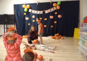 Dziewczynka leży na podłodze. Chłopczyk wraz z panią mierzy dziewczynkę za pomocą dyni.