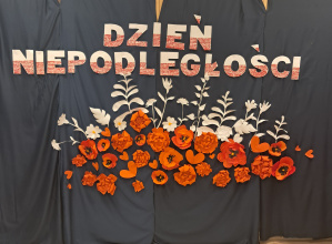 Dekoracja z napisem "Dzień Niepodległości" pod którym znajdują się białe i czerwone przestrzenne kwiaty.