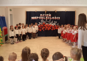 Dzieci z grupy Biedronek i Pszczółek stoją po dwóch stronach sali gimnastycznej na tle dekoracji. Pozostałe dzieci je obserwują.