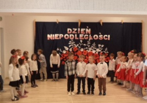 Dzieci z grupy Biedronek i Pszczółek stoją po dwóch stronach sali gimnastycznej na tle dekoracji. Czterech chłopców stoi na środku i recytyuje wiersze.