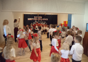 Dzieci ubrane na galowo i w barwy polskiej flagi stoją na sali gimnastycznej i obserują nauczycielkę.